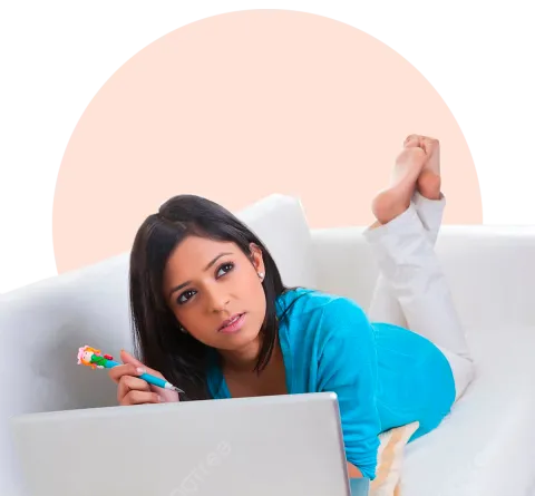 A woman lying on a white couch, thinking while holding a pen and looking at a laptop screen, representing an online mental therapy session.